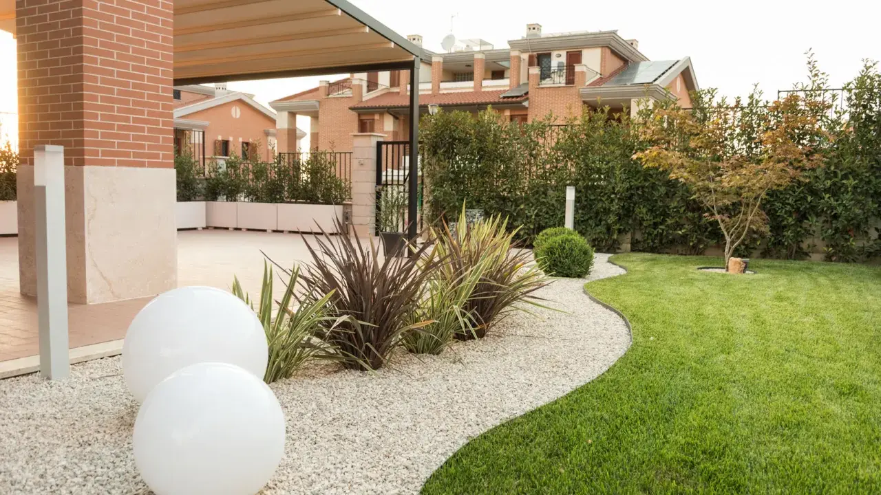 Aiuole davanti casa con ghiaia bianca, sfere luminose e prato verde curato. Sullo sfondo, case moderne e siepi.