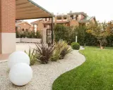 Aiuole davanti casa con ghiaia bianca, sfere luminose e prato verde curato. Sullo sfondo, case moderne e siepi.