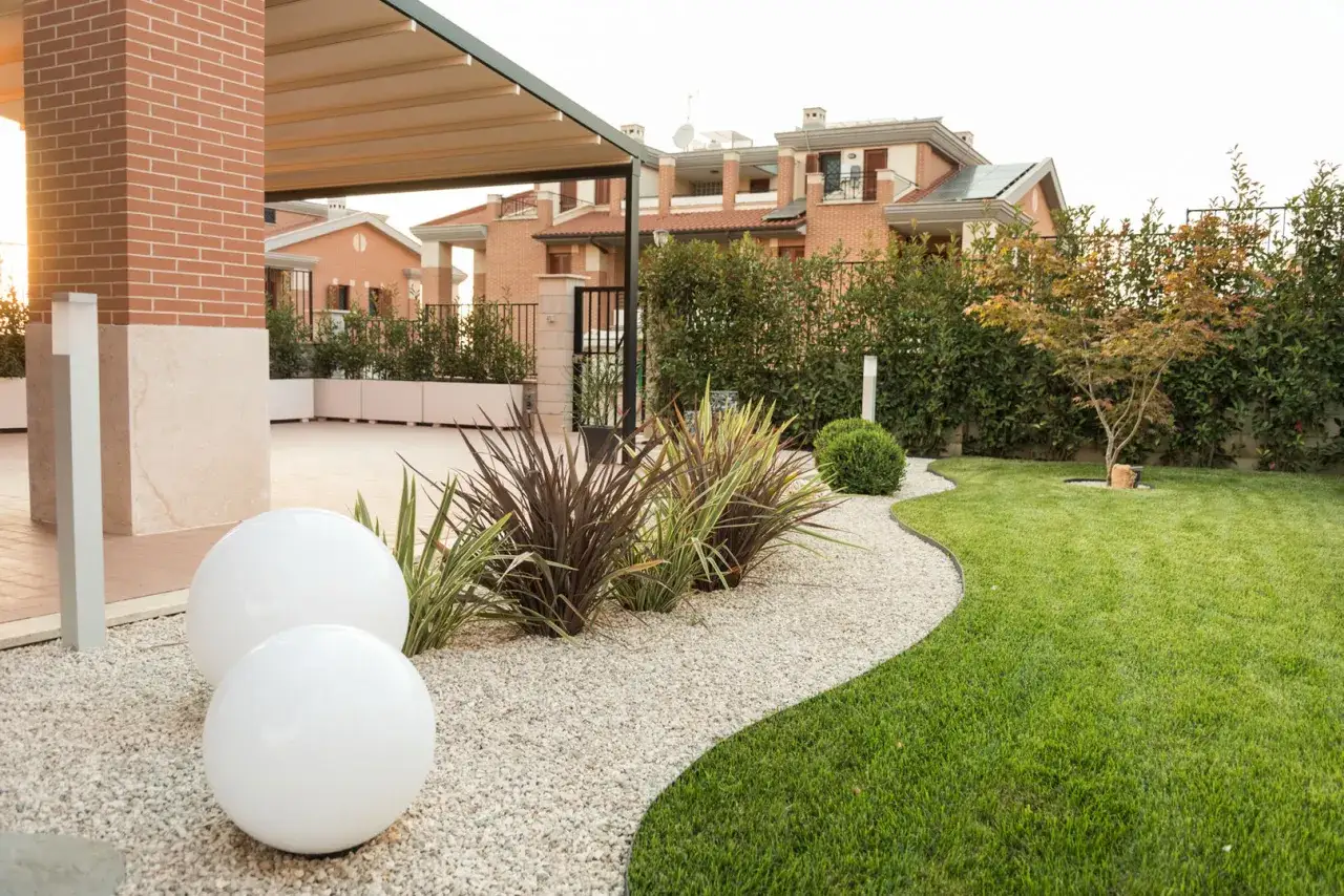 Aiuole davanti casa con ghiaia bianca, sfere luminose e prato verde curato. Sullo sfondo, case moderne e siepi.