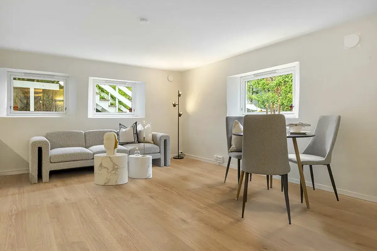 Soggiorno moderno con divano grigio, tavolini rotondi e zona pranzo. La casa beige e bianca è illuminata da finestre che mostrano il verde esterno.