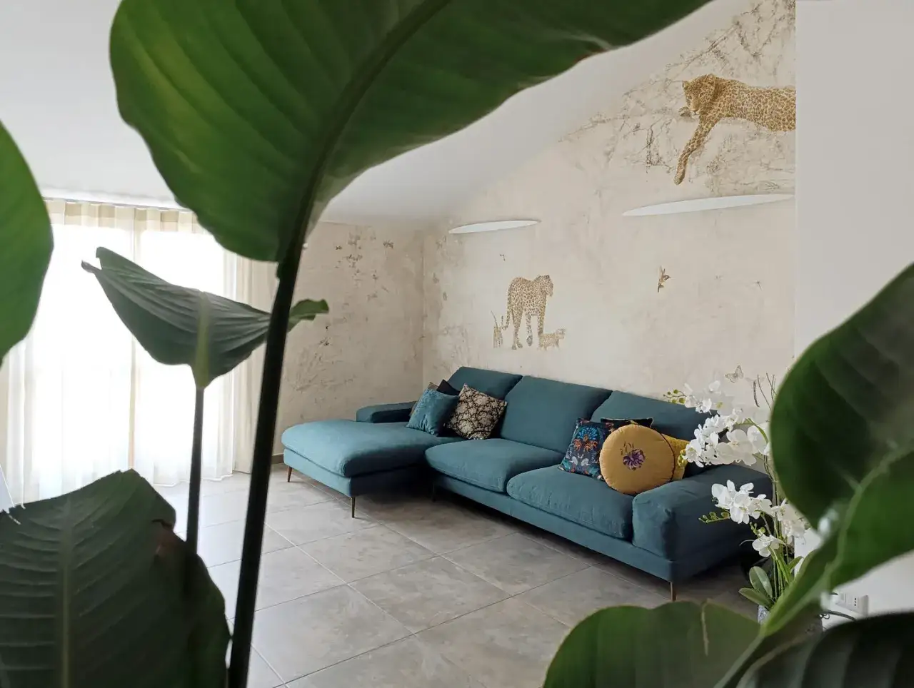 Divano turchese in un salotto con carta da parati a tema giungla e piante rigogliose, per un perfetto stile jungle chic.