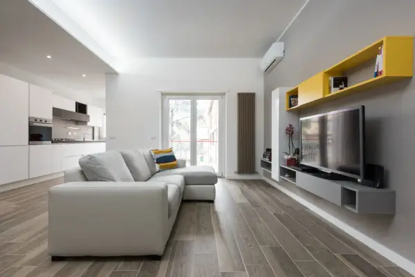 Un progetto soggiorno stretto e lungo con divano grigio, TV a parete e mensole gialle. La cucina moderna è visibile sullo sfondo.